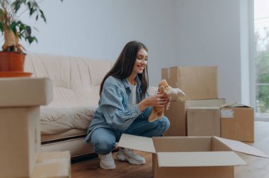 Neşeli genç bir kadın, eşyalarını rahat bir yaşam alanında kutuları taşırken gülümsüyor..