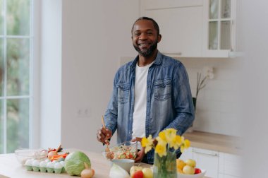 Neşeli bir adam, sebze, meyve ve çiçeklerle dolu, sağlıklı bir yaşam tarzının göstergesi olan, parlak, modern bir mutfakta taze bir salata hazırlıyor..