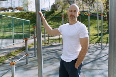 Kendine güvenen bir adam açık hava fitness parkında duruyor, yeşil, güneşli bir ortamda güç ve zindeliğin özünü sergiliyor..
