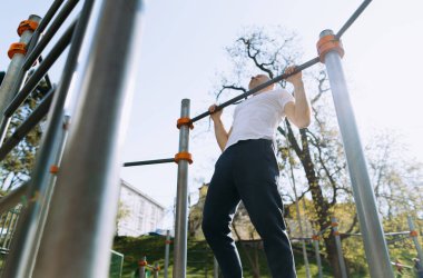 Bir adam güneşli bir havada parktaki barlarda barfiks çekerek egzersiz yapıyor. Güç ve zindelik gösterisi yapıyor..