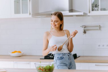 Beyaz kolsuz tişörtlü mutlu bir kadın modern bir mutfakta taze bir salata hazırlar. Sağlıklı bir yaşam tarzını benimserken ahşap bir kaşıkla yemek pişirmekten zevk alır..