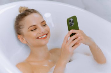 Köpük banyosunda gülümseyen bir kadın akıllı telefonunun keyfini çıkarıyor, rahatlık ve lüks bir ortamda modern özgüvenin vücut bulmuş hali..
