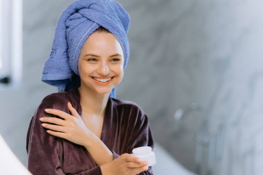 Mor bornoz ve mavi havluyla neşeli bir kadın cilt rutininin keyfini çıkarıyor. Hafif ışıklandırma ve mermer aksanlı şık bir banyo ortamında mutluluk saçıyor..