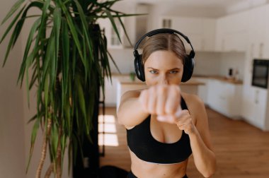 Odaklanmış bir kadın evde boks yapıyor, kulaklık takıyor ve şık bir iç mekanda güçlü yumruğunu sergiliyor..