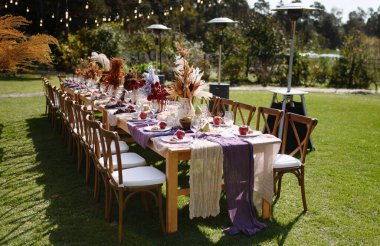 Çiçekler, meyveler ve sıcak aydınlatmalarla süslenmiş uzun bir masa içeren davetkar bir açık hava yemek düzeneği, toplanma için mükemmel..