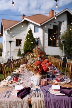 Zarif bir çiçek süsü, zarif bir cam süs eşyası ve dekoratif sicim lambası olan güzel bir açık hava yemek masası, büyüleyici bir evin arka planına kurulmuş..