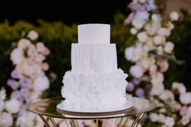 Zarif, beyaz bir düğün pastası, yumuşak pastel çiçeklerle çevrili dekoratif bir standın üzerinde duruyor. Romantik bir olay için mükemmel..