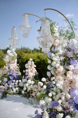 Göz kamaştırıcı bir çiçek aranjmanı üç parıldayan avizeyi çevreleyerek berrak mavi gökyüzünün altında canlı çiçeklerle dolu güzel bir açık hava ortamı yaratır..