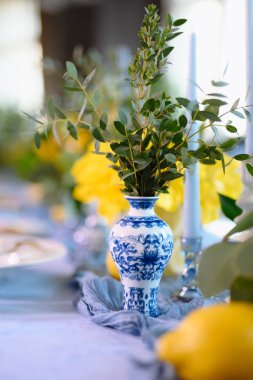 Şık bir masa sehpası, yeşil çiçeklerle ve mumlarla çevrili güzel mavi ve beyaz bir vazo sergiliyor..