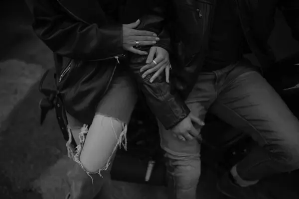 Bir çift hassas bir anı paylaşıyor, motosiklette oturuyor, şık deri kıyafetlerini sergiliyor ve birbirine dolanmış ellerini gösteriyor..