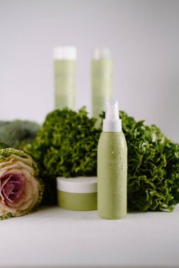 Taze yapraklı sebzelerle çevrili yeşil kozmetik şişeleri ve krema kavanozu..