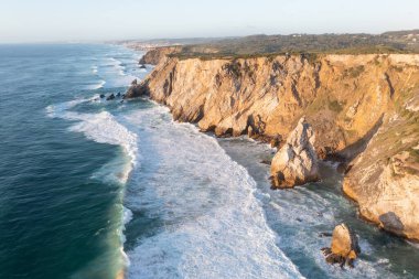 Atlantik Okyanusu 'nun Atlantik kıyısındaki Portekiz Ursa Sahili' nde kayalar ve günbatımı güneş dalgaları ve sahil şeridi manzaralı kumsalda köpükler. Praia da Ursa Sahili Sintra-Cascais Doğal Parkı