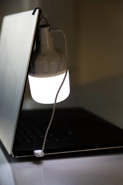 Portable rechargeable LED emergency light with built-in battery is charged from a laptop using a USB cable. Light source during blackout. Vertical shot.