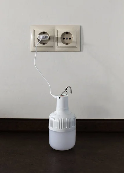 The portable rechargeable LED emergency light with built-in battery is charged from a 220V outlet using a USB cable and adapter. Light source during blackout. Vertical shot.