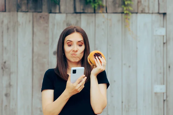 困惑的女人吃汉堡包检查她的手机 — 图库照片