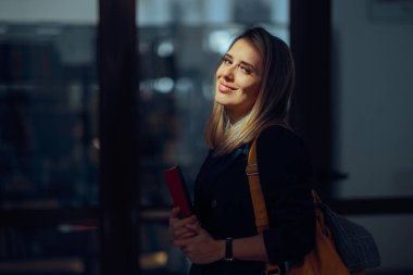 Gülümseyen Öğretmen Kütüphane önünde ders kitabı tutuyor.