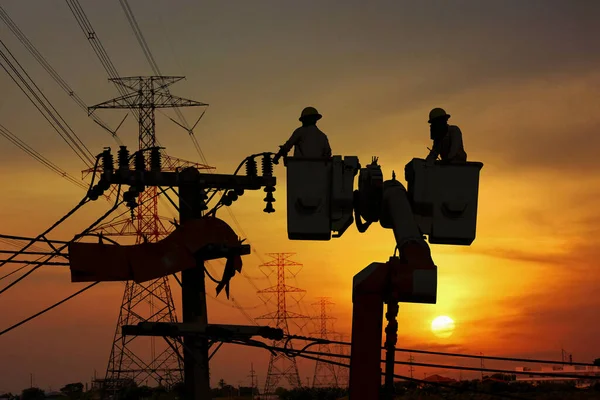 Bir grup siluet elektrik işçisi yüksek voltaj transformatörüyle yüksek voltaj iletim hattında çalışıyor.