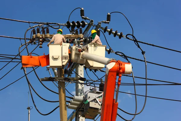electric engineering team working on cradle of aerial platform or crane connected high voltage transmission lines to high voltage transformer mounted on pole against blue sky