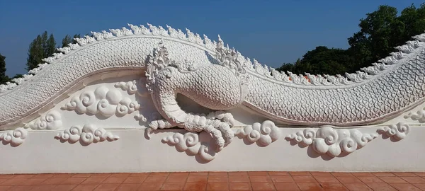 Three-dimensional Thai style stucco pattern on a white dragon\'s body, a wall of clouds contrasting with the sky.