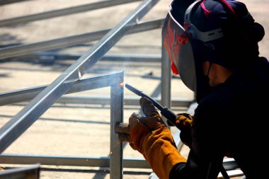 İnşaat alanındaki kaynakçı çelik yapısını kapatın. Kaynakçı deri eldiven ve kask giyerken işçi kaynakçıları elektriği ve sıcaklığı korur.