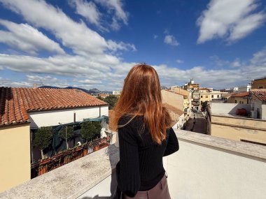 Yatay geniş açılı fotoğraf, kırmızı saçlı bir kadının çatı terasında dikiz görüntüsünü gösteriyor. Olbias 'ın eski kasabası Sardinya' nın panoramik manzarasına hayran..