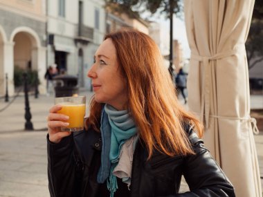 Siyah ceketli genç kızıl saçlı kadın ve mavi atkı Piazza Matteotti 'deki açık kafede taze portakal suyunun tadını çıkarıyor, Olbia Old Town, Sardinya