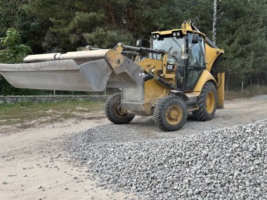 Bir kazıcı bir kovayla ezilmiş taşı topluyor. Traktör toprak bir yolu dümdüz eder. Yol çalışması, orman yolunun yakınındaki makineler. Ukrayna, Kiev bölgesi, Irpen şehri, 12 Ekim 2023