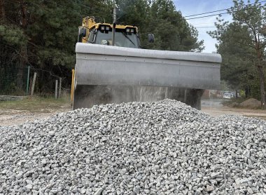 Bir kazıcı bir kovayla ezilmiş taşı topluyor. Traktör toprak bir yolu dümdüz eder. Yol çalışmaları, orman yolunun yakınındaki makineler..