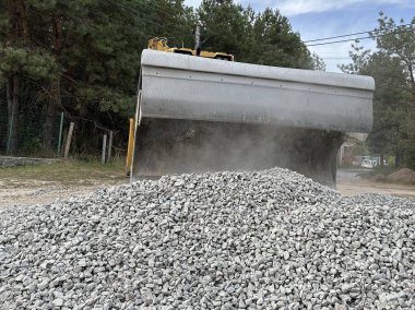 Bir kazıcı bir kovayla ezilmiş taşı topluyor. Traktör toprak bir yolu dümdüz eder. Yol çalışmaları, orman yolunun yakınındaki makineler..