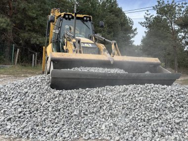 Bir kazıcı bir kovayla ezilmiş taşı topluyor. Traktör toprak bir yolu dümdüz eder. Yol çalışması, orman yolunun yakınındaki makineler. Ukrayna, Kiev bölgesi, Irpen şehri, 12 Ekim 2023