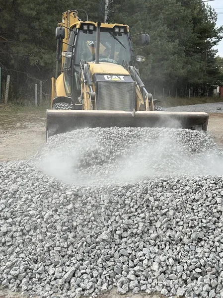 Bir kazıcı bir kovayla ezilmiş taşı topluyor. Traktör toprak bir yolu dümdüz eder. Yol çalışması, orman yolunun yakınındaki makineler. Ukrayna, Kiev bölgesi, Irpen şehri, 12 Ekim 2023