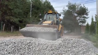 Bir kazıcı bir kovayla ezilmiş taşı topluyor. Traktör toprak bir yolu dümdüz eder. Yol çalışması, orman yolunun yakınındaki makineler. Ukrayna, Kiev bölgesi, Irpen şehri, 12 Ekim 2023
