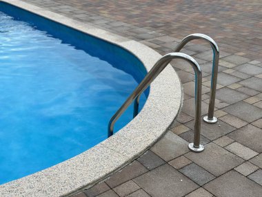 Giriş için tırabzanları olan yüzme havuzu, yakın plan. Havuzda şeffaf mavi su ve paslanmaz çelik parmaklıklar..