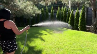 Yeşil çimenleri hortumla sulayan bir kadın. Çimlere ve katil geçmişine karşı yeşil bahçeyi sulamak. Su güneşe ve bahçedeki yeşil bitkilere sıçrar.