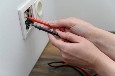 female hands use the probes of a digital multimeter to check the current voltage in an electrical outlet.
