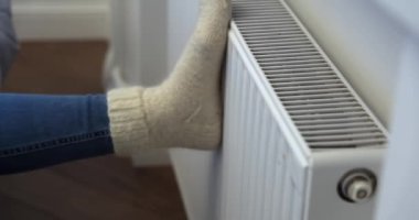 Beyaz yün çoraplı bir kadın kış boyunca ısıtma kaloriferinin önünde soğuk ayaklarını ısıtıyor. Evde elektrikli ya da gazlı ısıtıcı var..