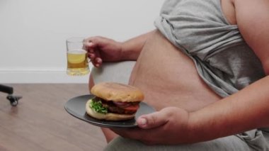 Hamburger ve bir bardak birayla şişman bir adam televizyon izler..