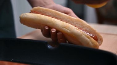 Modern bir mutfak ortamında ızgara sosisliye hardal sosu ekleyen bir elin yakın çekimi.