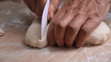 Sıcacık bir mutfak ortamındaki ahşap bir tezgahta ustalıkla taze hamuru bölen zanaatkar ellerinin yakın çekimi.