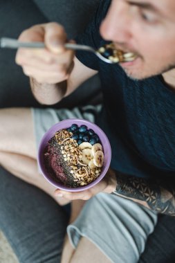 Adam kahvaltıda müsli ve meyve ve fındık yerken kanepede oturuyor. Dikey fotoğrafçılık. Seçici odaklanma. Sağlıklı yaşam tarzı kavramı