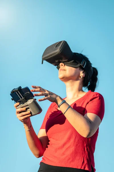Latin woman flying drone with virtual reality fpv headset in outdoors blue sky on a sunny day. Vertical photography