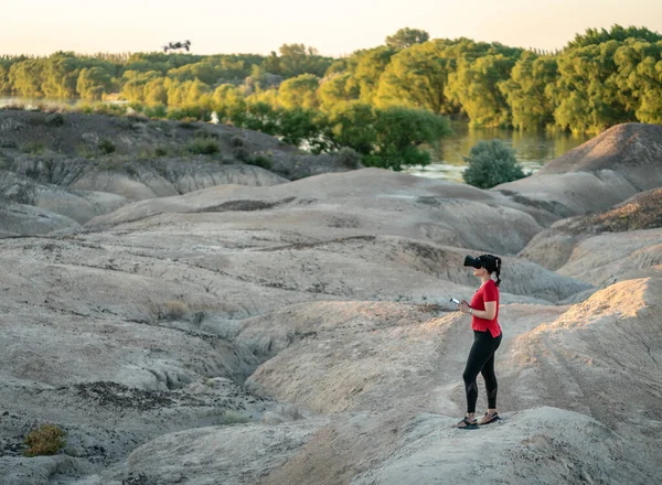 Woman flying fpv drone outdoors while using virtual reality headset in arid badlands area. Panoramic view