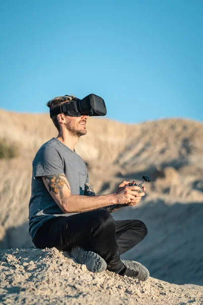 Man sitting in arid outdoors area flying drone with virtual reality mask. Vertical photography