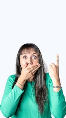 Surprised young Latin woman covering her mouth with one hand and pointing above with the other one. Isolated white background with copy space for advertisement