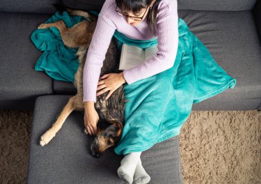 Top view of woman petting her crossbreed dog while reading a book on the couch. Relax time during cozy winter