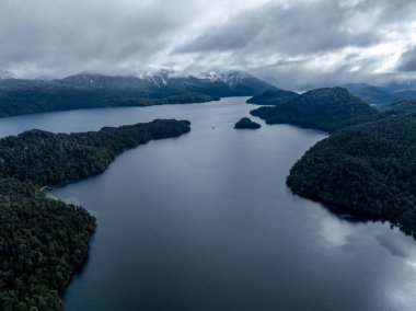 Yoğun bir orman ve bulutlu bir gökyüzünün altındaki sis kaplı dağlarla çevrili bir göldeki adanın havadan görünüşü.