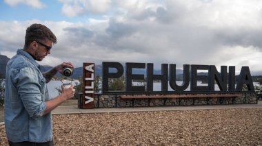 Villa Pehuenia Köyü 'nün karşılama tabelasının önünde içkiye su döken adam. Patagonya 'da bir turizm merkezi. 4 Nisan 2025. Villa Pehuania, Neuquen, Arjantin