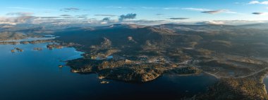 Havadan gelen panoramik, Alumine Gölü 'nü birkaç adayla, kısmen bulutlu bir gökyüzünün altında orman tepeleri ve dağlarla çevrili olarak gösteriyor. Villa Pehuenia, Neuquen, Arjantin