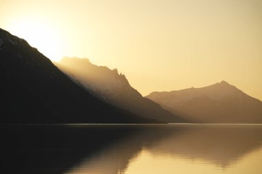 Huzurlu göl kenarındaki dağ tepelerinin silueti ile destansı altın gün batımı. Şaşırtıcı doğal manzara.