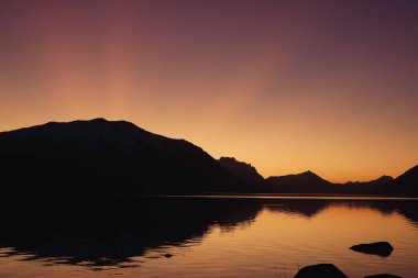 Gölün üzerinde dağların siluetleri olan renkli bir alaca karanlık.